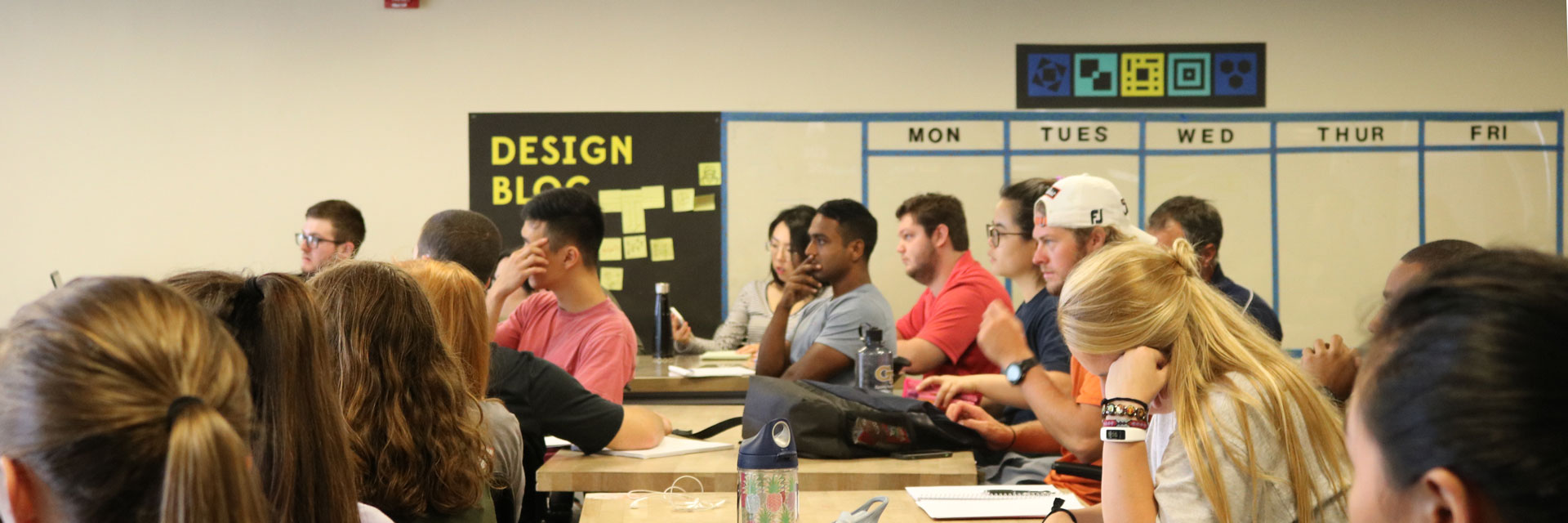 An audience of students, viewed from the side, listens intently to a Design Bloc professor at the front of the class.
