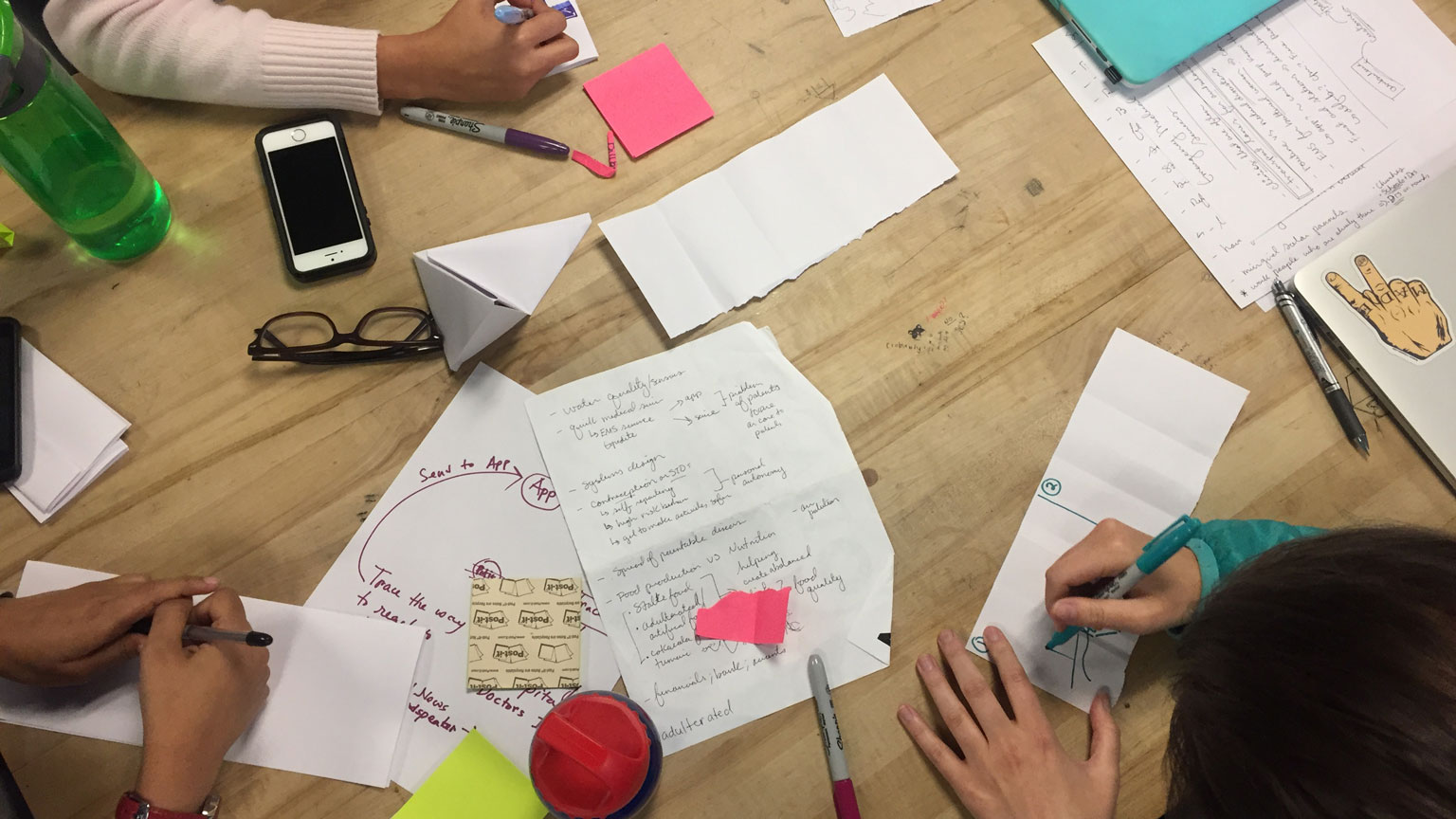 Hands surround a table full of post-it notes and sketches during a brainstorming session. 
