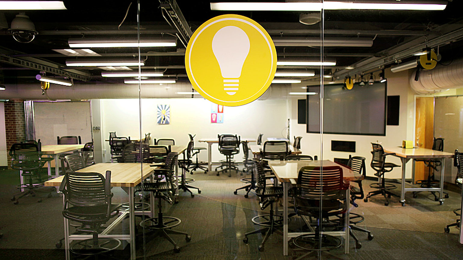View into a classroom full of rolling tables and chairs, a presentation screen in the background, and a large lightbulb symbol hanging from the glass wall in center-view.. 